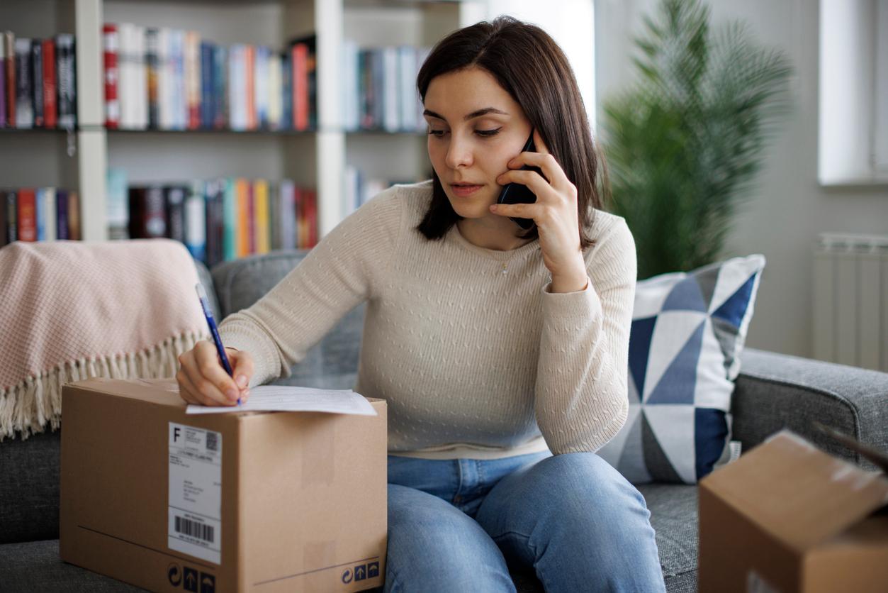 A woman sitting on a sofa, talking on the phone while writing on a document placed on a cardboard package.