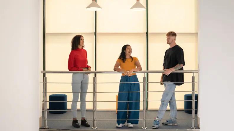 Three coworkers having a conversation near a railing in a modern office setting, fostering collaboration.