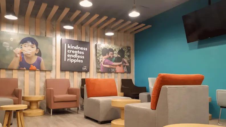 A cozy office lounge with warm lighting, modern seating, and wooden panel decor. The walls feature motivational artwork, including a sign that reads 'Kindness creates endless ripples.