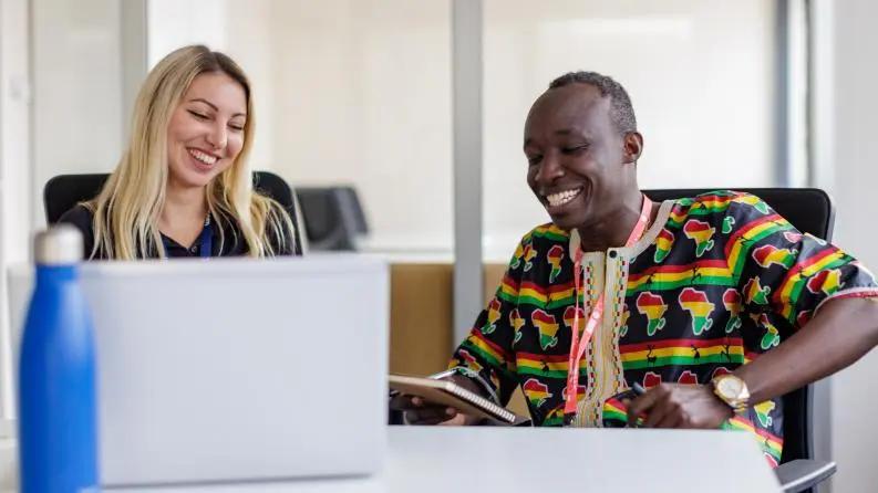 Two colleagues smiling and discussing work at an office desk with a laptop