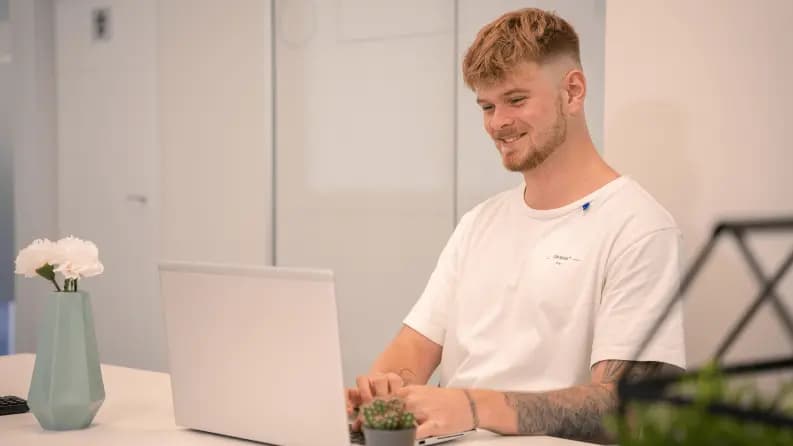 Smiling man in a white t-shirt working on a laptop in a bright office.