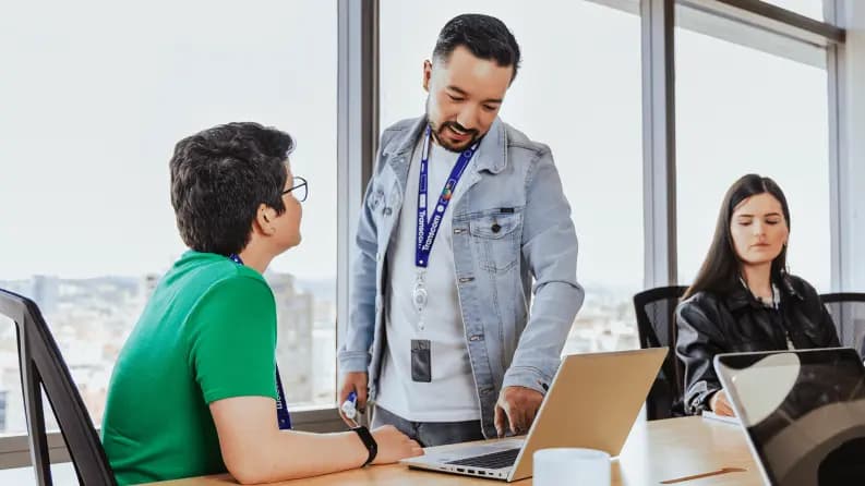 Three colleagues in an office, one standing and discussing something on a laptop while the others listen
