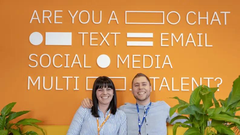 Two employees smiling in front of an orange wall with text and symbols, representing a friendly workplace environment.