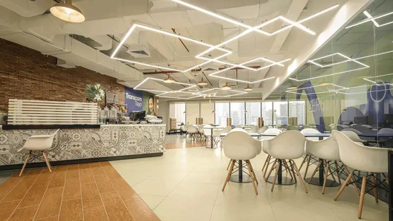 A stylish office cafeteria with modern white chairs, wooden and tiled flooring, a patterned counter, and geometric LED ceiling lights. The space has a mix of open seating and a glass-walled meeting room.