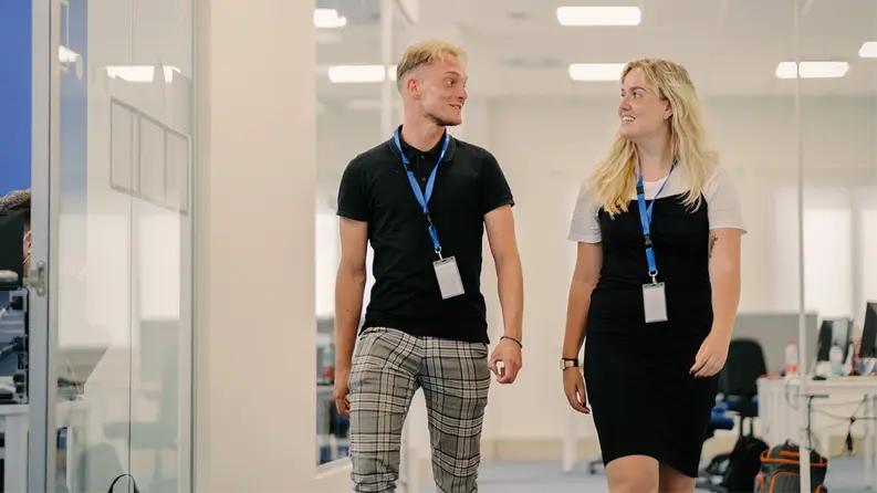 Two employees smiling in front of an orange wall with text and symbols, representing a friendly workplace environment.
