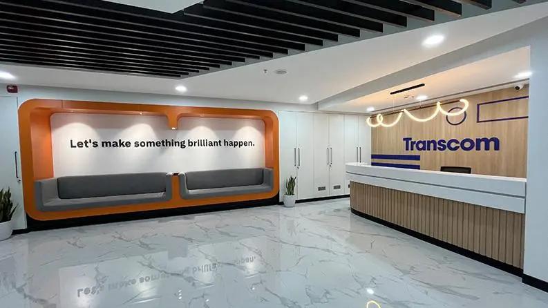 Sleek office reception area with a Transcom-branded front desk and a seating area featuring the quote 'Let's make something brilliant happen.