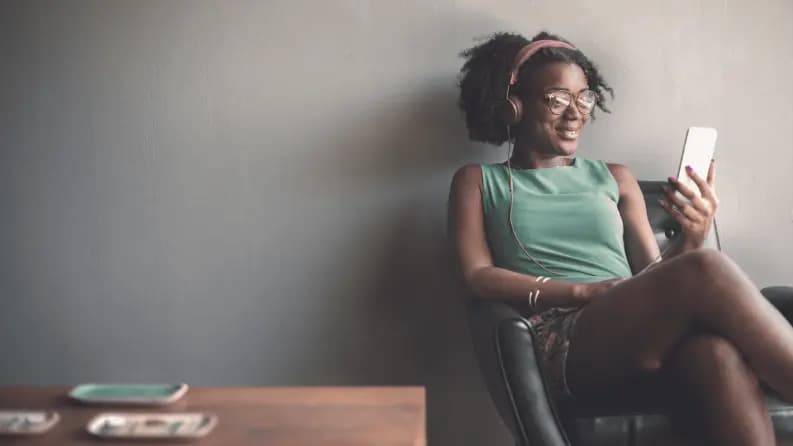 A woman sitting in a chair, wearing headphones and smiling while looking at her smartphone.